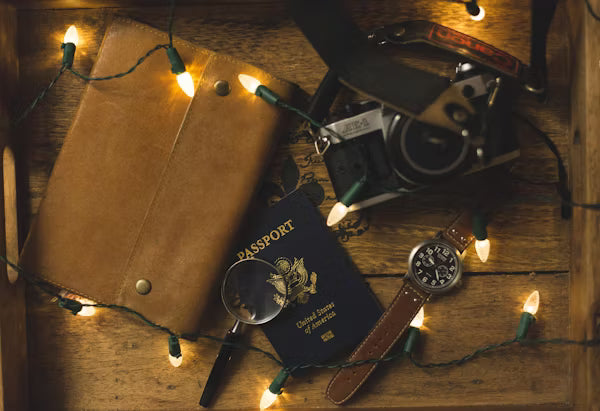 Vintage camera, passport, magnifying glass, and leather wallet on a wooden surface with string lights.