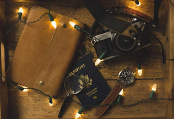 Vintage camera, passport, magnifying glass, and leather wallet on a wooden surface with string lights.